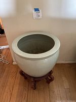 Single image showing a large light green ceramic planter on a carved wooden stand on a hardwood floor against a white wall.
