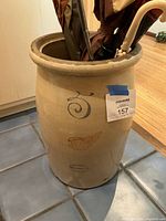 Ceramic umbrella stand with several umbrellas stored inside. Beige stoneware with blue stamped '5' and brown wing design. Lot tag attached.