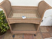 Full view of garden trolley showing faux wicker construction, two shelves with glass tops, curved handles on sides, and caster wheels.