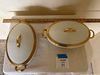 Top view of two oval ceramic covered dishes with gold detailing and handles, alongside a ruler for size reference.