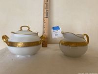 Front view of sugar bowl with lid and cream pitcher with gold ornate band and handles alongside a ruler for size.