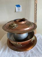 Copper Mongolian hot pot with lid slightly open revealing inner pot; placed next to a ruler for size reference.