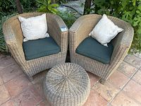 Two wicker armchairs with cushions and round ottoman arranged outside on a brick patio with greenery background.