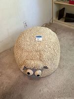 A sheep-shaped plush footstool with shaggy cream fabric and large cartoon-like eyes and felt ears, placed on a carpeted floor near a wall outlet.