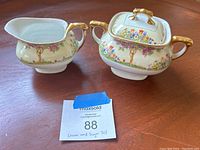 Cream pitcher and sugar bowl set on wooden surface showing floral design and gold trim.