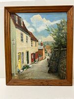 Full view of the painting showing a village street scene with houses and greenery in a wooden frame.