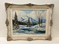 Framed oil on board painting showing entire winter scene with snow-covered mountains, pine and bare trees, and a river.