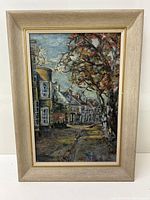 Front view of framed oil on board painting depicting a village street scene with buildings and large tree.