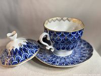 Porcelain teacup with saucer and detached lid, showing intricate blue and gold Forget Me Not pattern.