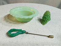 Lot contents showing jadeite bowl, grape cluster figurine, twisted jade pin with gold-tone fastening, and green plastic ring on cloth background.