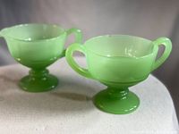 Photo showing two light green jadeite glass pieces: a creamer on the left and an open sugar bowl with two handles on the right, both with pedestal bases.