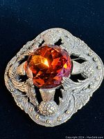 Clear close up of the brooch front showing large central amber-colored faceted gemstone and detailed thistle and heart motifs in silver-tone metal.