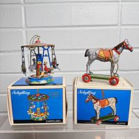 Both Schilling tin toys shown on top of their original boxes against a white brick wall background. Carousel is colorful with figures on swings. Horse is gray with polka dots and mounted on red wheels.