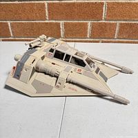 Side angle of beige Star Wars SnowSpeeder toy showing detailed front cannons, cockpit, and rear turret against brick background.