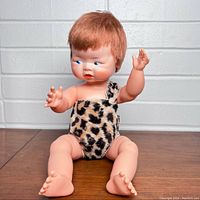 Front angled view of the Pebbles Flintstone doll sitting on a wooden surface against a white brick background. Shows the doll's facial details, hair, and one-shoulder leopard outfit.