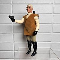 Full view of the Battlestar Galactica figure standing upright against an off-white textured wall; holding a black toy gun, dressed in beige outfit with brown vest and black boots.