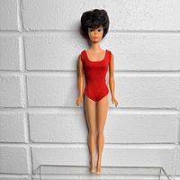 Front full-length view of vintage Barbie doll in red swimsuit against white brick background, showing tight bubblecut dark brown hair and detailed facial paint.