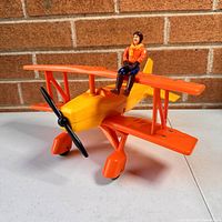 Front side view of the bright orange and yellow biplane with seated toy figure on top wing.