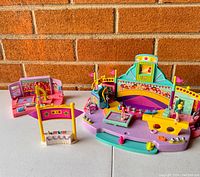 Two Polly Pocket gymnastics-themed playsets shown on a flat surface against a brick wall background. Includes a gymnastics stage with female figurines, audience seating, and apparatus, plus a smaller gymnastics equipment set.