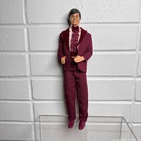 Full body view of vintage Ken doll in burgundy tuxedo against white brick background.