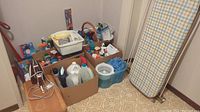 Various cleaning supplies in boxes including large plastic bottles of cleaning liquids, an iron, a white plastic bin with cloths, and mop head in blue bucket alongside an ironing board.