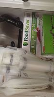 Photo showing FoodSaver brand vacuum storage bag boxes and rolls stacked beside a kitchen sink faucet.