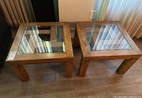 Two square side tables made of wood with thick glass tops shown from above on wood floor near window with blinds.