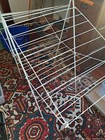 Full view of the white metal vintage laundry rack fully opened and standing on a patterned carpet, showing multiple horizontal bars for hanging clothes.