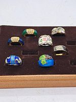 View of all 7 assorted vintage rings in a display tray, showing various colored enamel, rhinestone and floral designs.