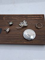 Five pieces of sterling silver jewelry set on a wooden board, including a floral brooch, round brooch, and three rings.