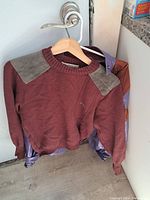 Full view of burgundy wool sweater with beige suede shoulder patches hanging on a hanger with purple leather jacket behind it.