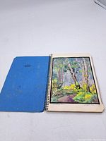 Open spiral-bound blue-covered book showing a full-page landscape watercolour painting with trees and a pathway.
