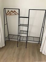 Photo showing a modern black metal clothes rack with three central grid shelves and two horizontal bars on each side for hanging clothes, placed against a wall on a wood floor.