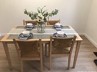 Dining table set with glass top and four wood and wicker chairs, set with blue and white dishes for scale