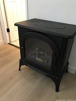 Black small electric fireplace with glass front and broken door handle on right side, standing on wooden floor near white wall.
