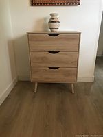 Front view of the small dresser showing three drawers and tapered legs in a light wood finish with a decorative vase on the top (vase not included in sale).