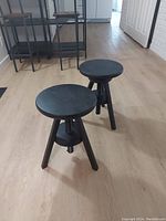 Two small black painted wooden stools with three legs and adjustable height mechanism. Positioned on floor in natural lighting.