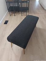 Full view of the black upholstered bench with gold legs placed on light wood floor.