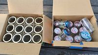 Top-view photo of two open cardboard boxes, each filled with empty glass canning jars. Left box shows 12 jars with metal screw rings but no lids. Right box contains 12 jars with metal lids featuring various designs and colors.