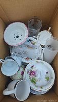 Box with various china cups, saucers, plates, clear and patterned glassware, and a flower pot.