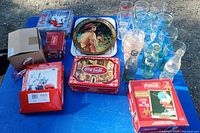 Coca-Cola collection displayed on blue tarp including metal tins, several drinking glasses, glass bottles, and boxed jigsaw puzzle.