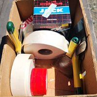 Box containing drywall tools including drywall knives, rolls of tape, and boxed hydraulic jack.