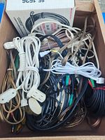 Box containing a variety of electrical cords and cables including white, black, clear, and brown cords with different plugs.