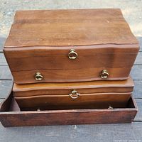 Wooden jewelry boxes stacked; top box has brass ring pull handles and natural wood finish. The bottom box has its drawer partially open, showing a similar design and hardware, set inside a rectangular wooden tray with a slightly darker finish.