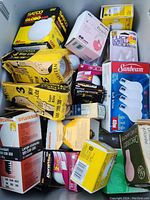 Multiple light bulbs of varying sizes and wattage in original boxes, from assorted brands packed in a plastic bin. Box colors include yellow, white, pink, black, green.