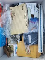 Items in a box including a Mead spiral notebook, rulers, pens, pencils, plastic wrapped items, and cardboard drawer organizer with assorted office supplies.