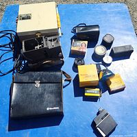 Top-down photo of vintage camera equipment laid out on a blue surface showing a Gaf Super Slide Projector, assorted lenses, film canisters, boxes of flash bulbs, and carrying cases.