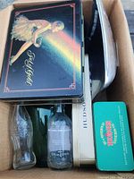 Box with stacked collectible tins: black tin with ballerina design, a Hudson Bay tin, and other tins; vintage glass bottles including Coca-Cola and I-Pop bottles partly visible