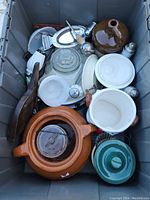 Overhead view of bin containing brown ceramic cookie jar, white bowls and plates, clear glass jars, small decor items, and salt and pepper shakers