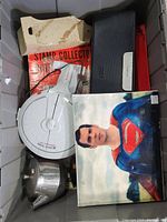 Photo showing a stamp collector kit with a book, a silver metal teapot, a Superman themed item, red and black boxes or cases, and a circular Star Trek Enterprise themed object.